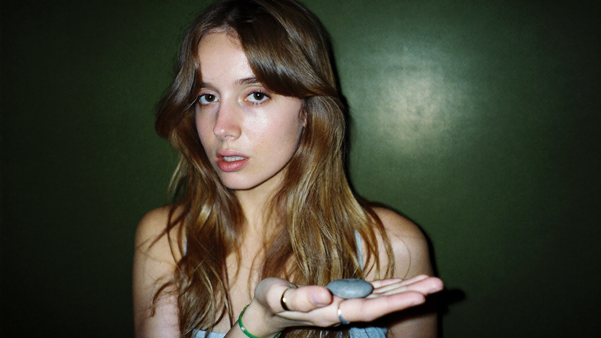 A young woman holds a smooth grey pebble in her open palm against a deep green wall