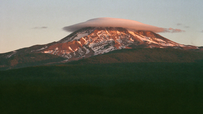 Mount Shasta at dusk with a lenticular cloud over its snowy summit above dark conifer forest