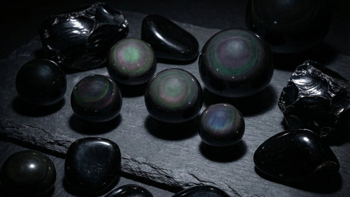 Polished rainbow obsidian spheres with green and violet sheen beside rough and tumbled black obsidian on dark slate