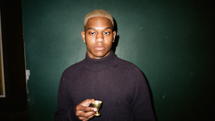 A young man in a dark turtleneck holds a small brass offering cup against a deep green wall