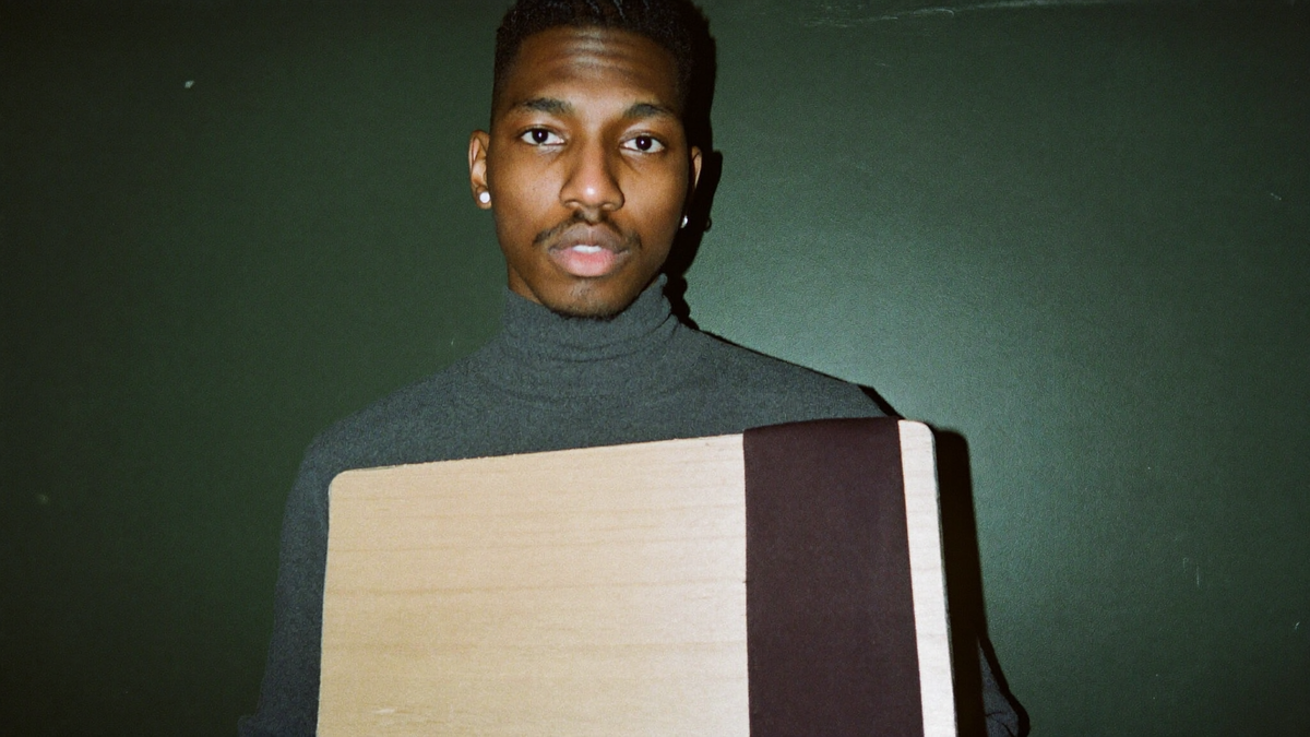 A young man in a grey turtleneck holding a plain wooden board against his chest, against a dark green wall
