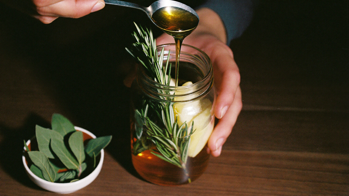 Honey poured from a spoon into a mason jar of rosemary, garlic and vinegar, with a bowl of fresh sage leaves beside it