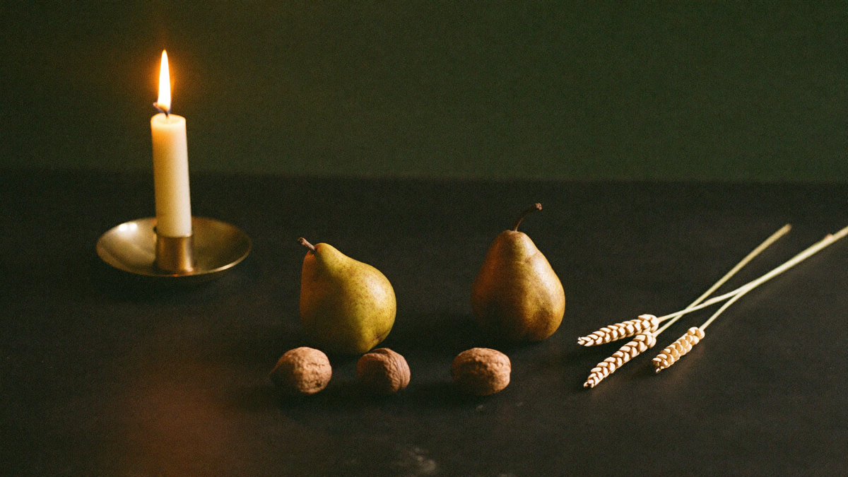 Two pears, walnuts and dried wheat stalks beside a single burning candle in a brass holder on dark stone