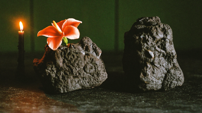Two rough black lava rocks with a single red hibiscus flower beside a burning candle on dark stone