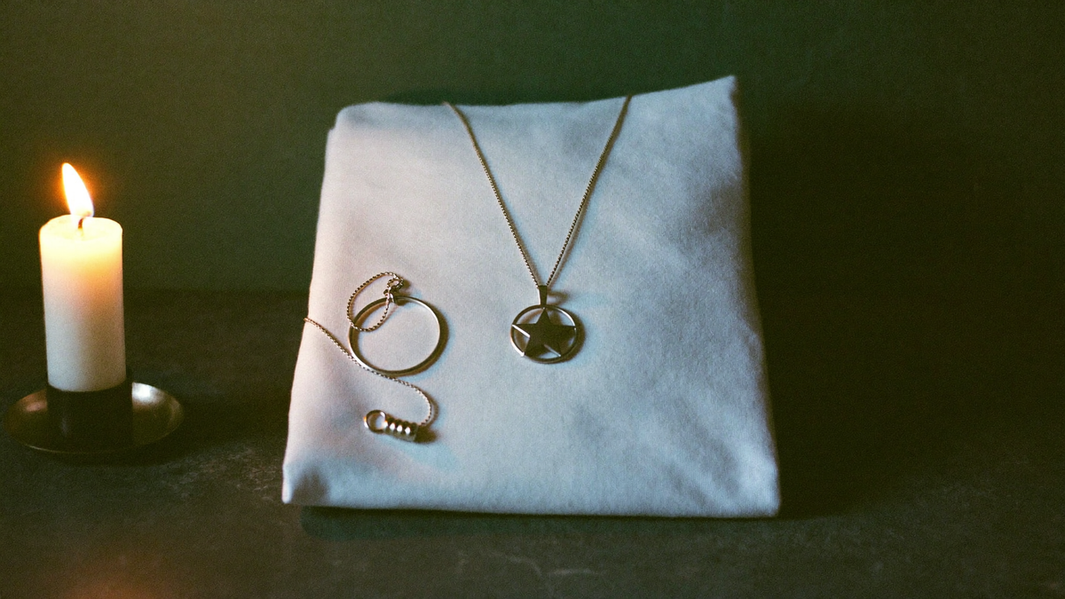 A silver pentacle pendant on a chain beside a folded white shirt, lit by a candle on dark stone