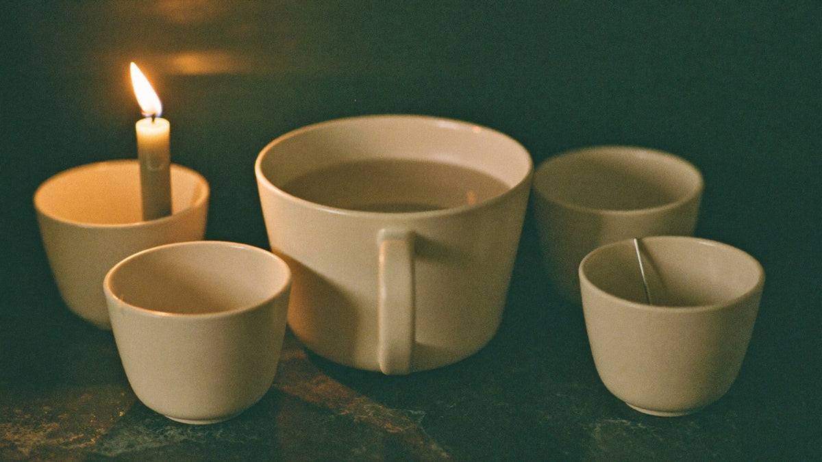 A full cream-colored ceramic mug surrounded by four small empty cups on dark stone, a lit taper candle on the left