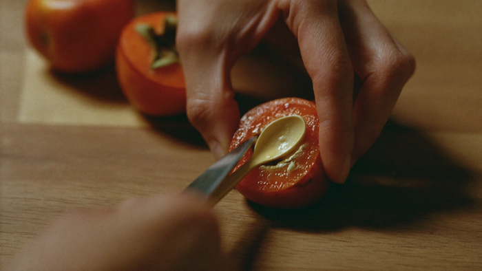 Two hands opening a ripe persimmon on a wooden board with a paring knife, the pale spoon shape of the seed showing, by candlelight