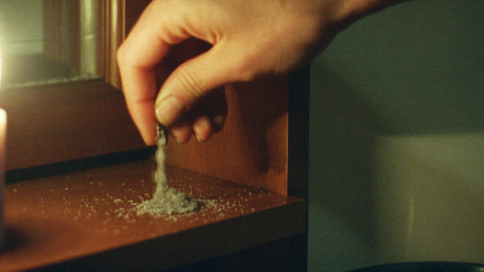 A hand pouring a thin line of coarse dark salt along a wooden threshold in warm candlelight