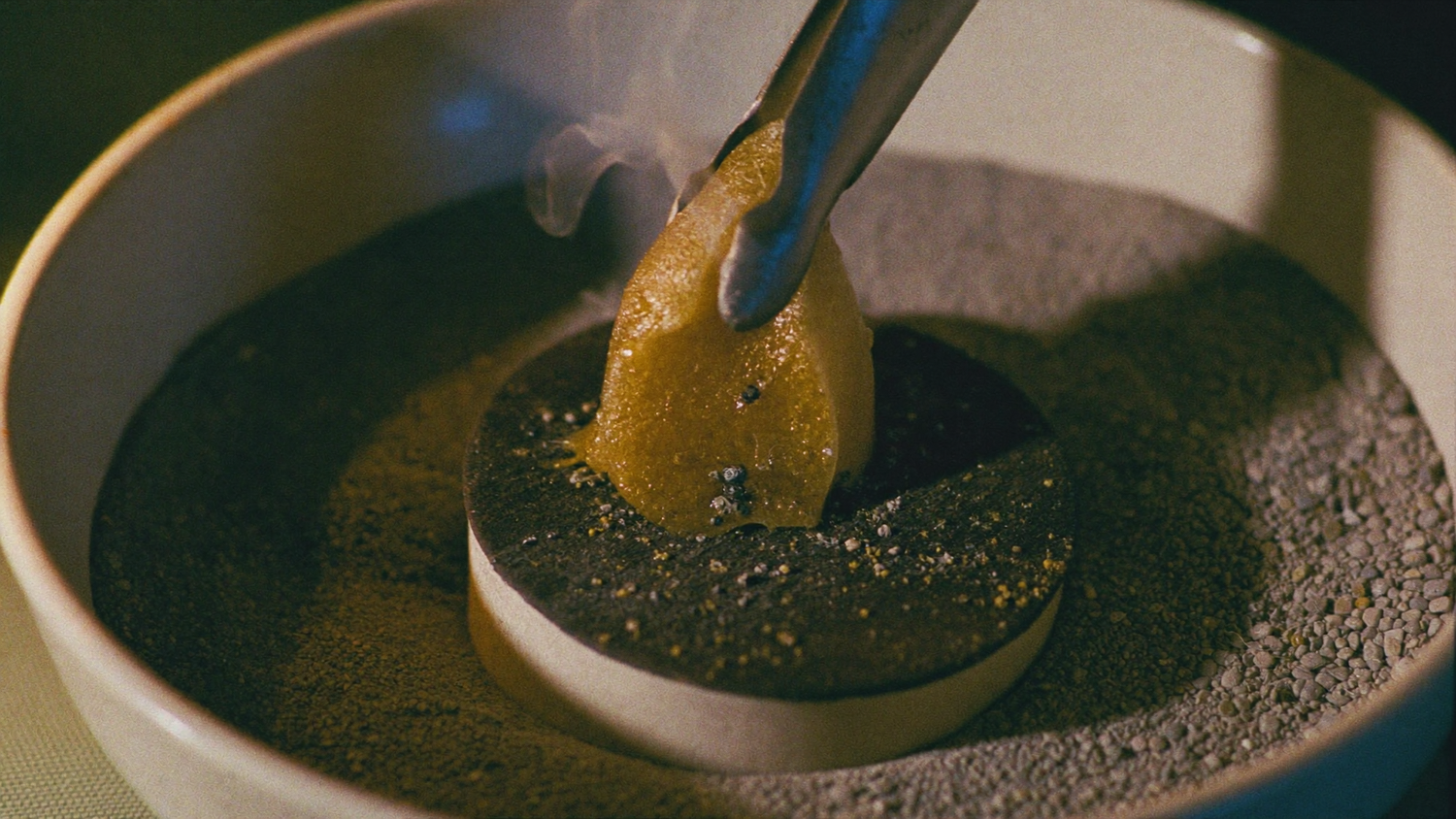 Metal tongs placing a lump of golden frankincense resin onto a charcoal disc set in sand inside a ceramic burner