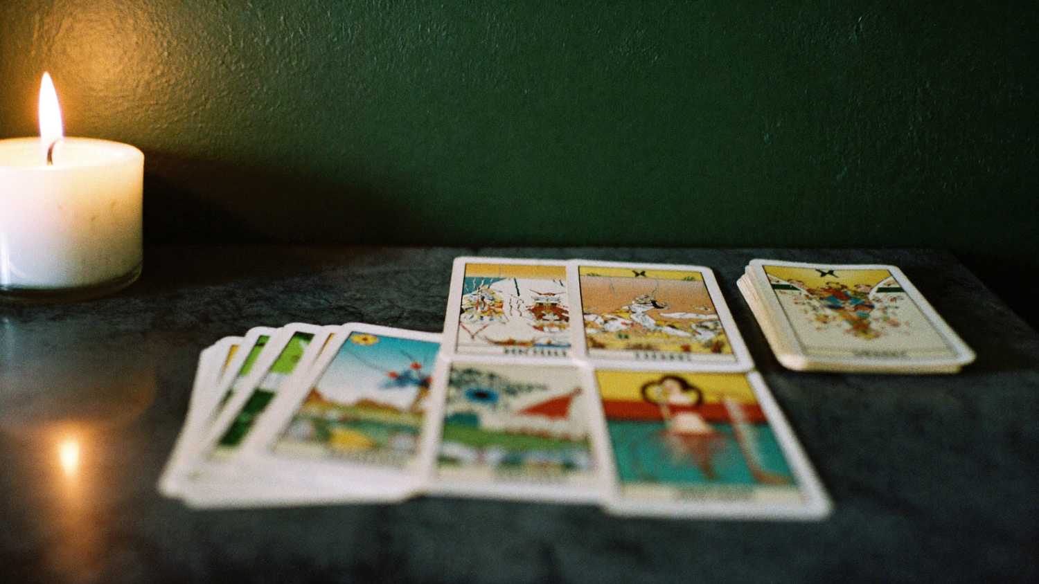 A worn tarot deck separated into small stacks on a dark stone table beside a burning candle