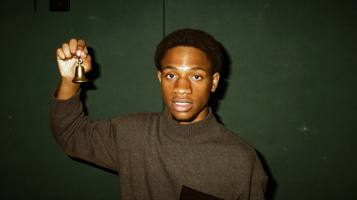 A young man in a turtleneck holding up a small brass hand bell against a dark green wall