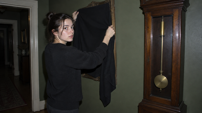 Woman draping a black cloth over a hallway mirror beside a grandfather clock with its pendulum stopped