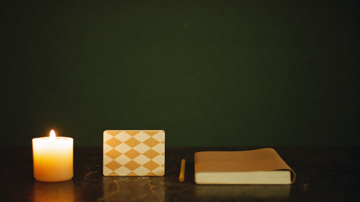A lit candle, a face-down stack of patterned cards, a pencil and a closed notebook on dark stone against a green wall
