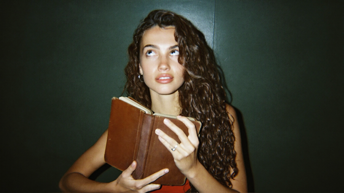 A young woman holding a worn leather book, looking up and away from the page in warm light