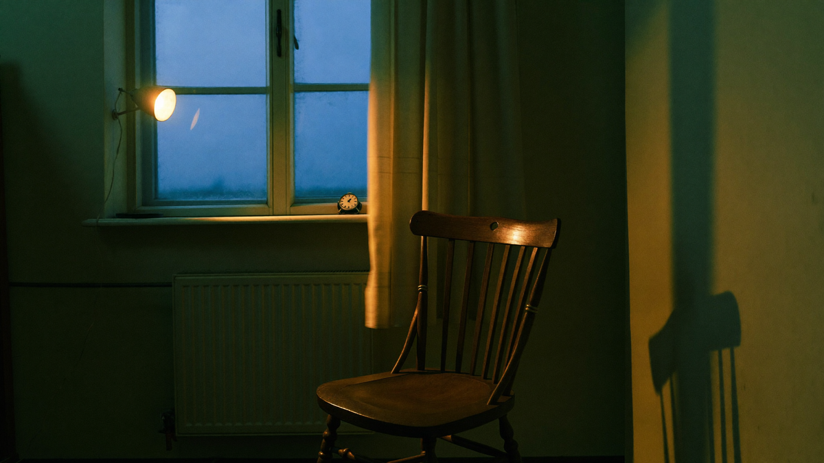 An empty wooden chair beside a window at blue hour, a small lamp lit and a clock on the sill