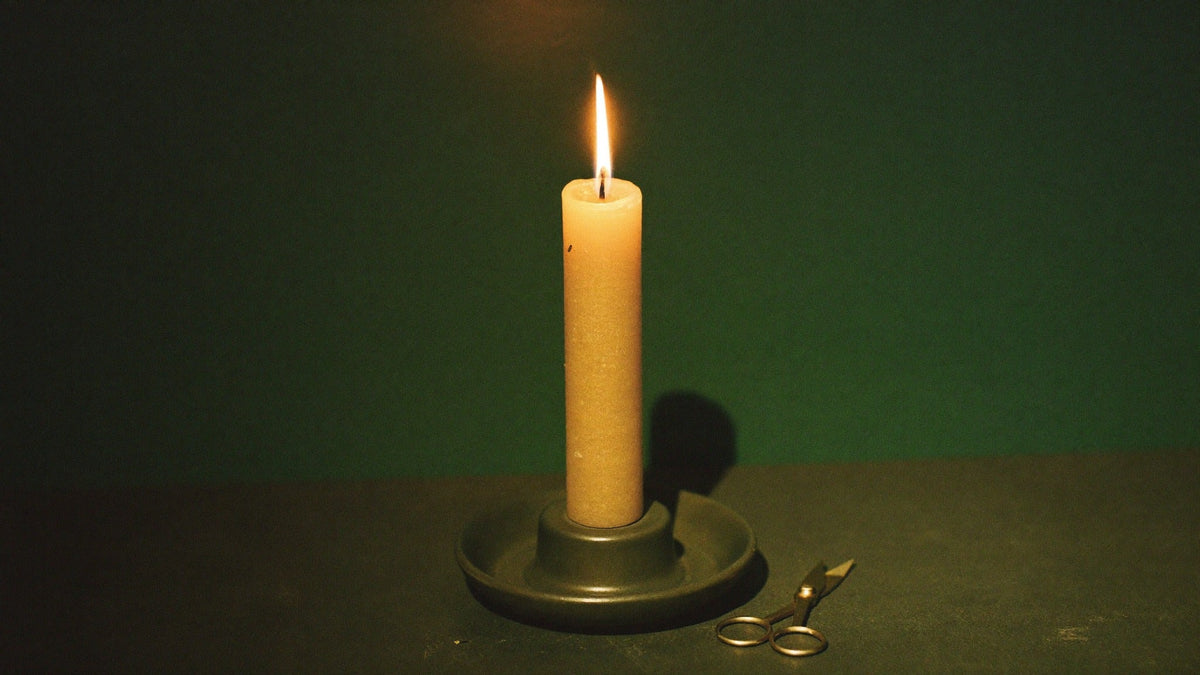 Lit beeswax pillar candle in a dark ceramic holder beside small brass wick trimmers against a deep green wall