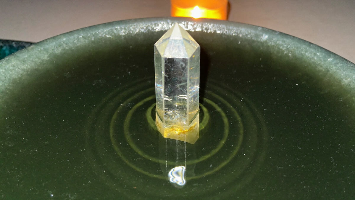 A clear quartz point standing upright in a shallow green bowl of still water, one ripple ring around its base