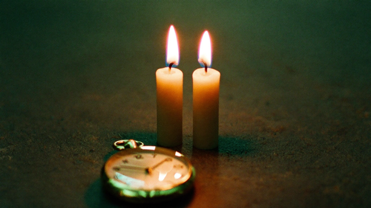 Two lit candles beside a gold pocket watch on a dark table, warm light in a quiet green room