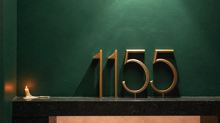 Brass house numbers 1155 mounted on a dark green wall above a black stone ledge with a small burning candle