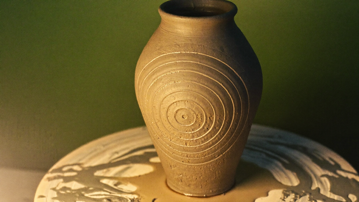 Freshly thrown clay vessel standing on a potter's wheel head, spiral throwing rings visible in the soft grey clay