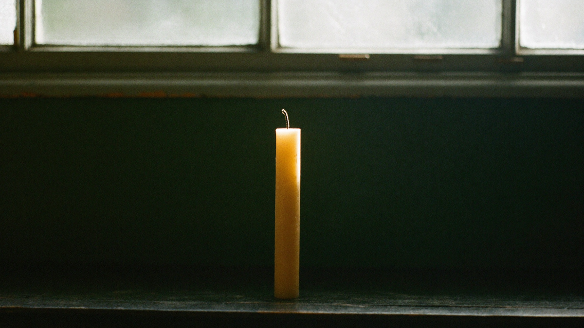 Tall unlit beeswax candle with an unburned wick standing on a worn dark windowsill in cold first daylight