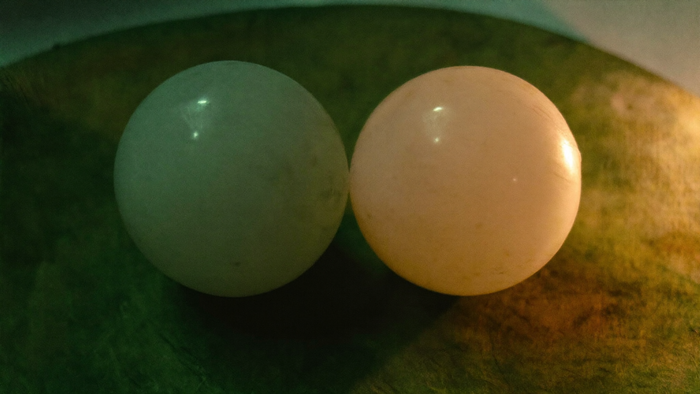 Two pale moonstone spheres resting side by side in a dark green stone dish, one lit warm, one cool
