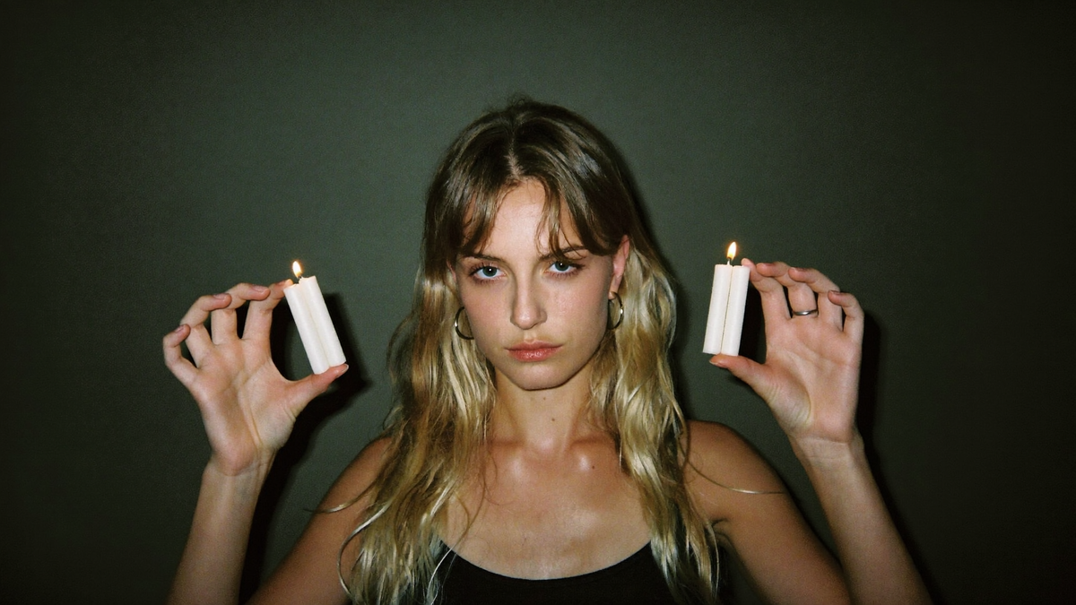 A young woman facing the camera holding two lit white candles in each hand against a dark green wall