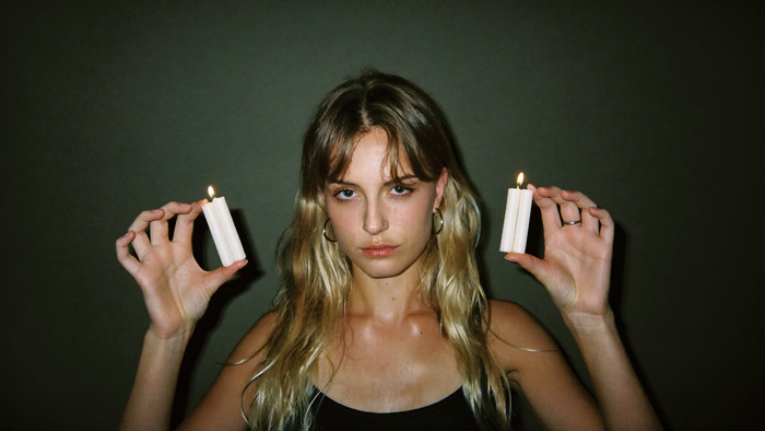 A young woman facing the camera holding two lit white candles in each hand against a dark green wall