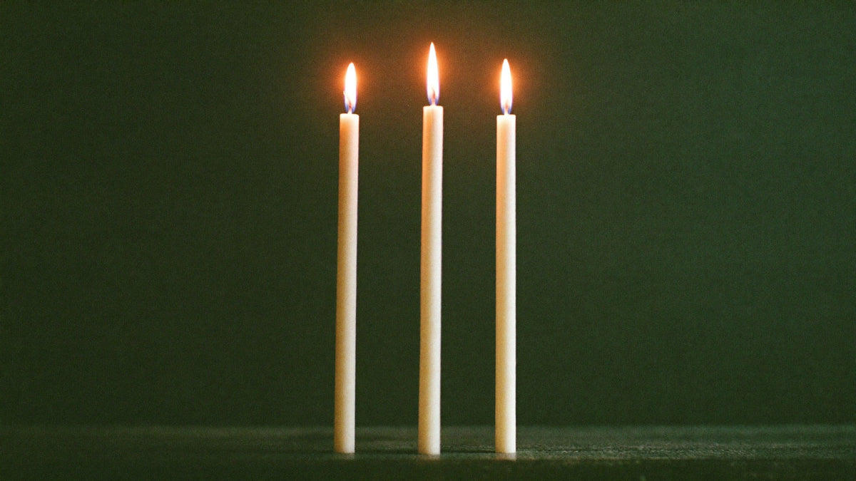 Three slender white candles burning side by side against a dark green wall, a symbol of the three in 3333