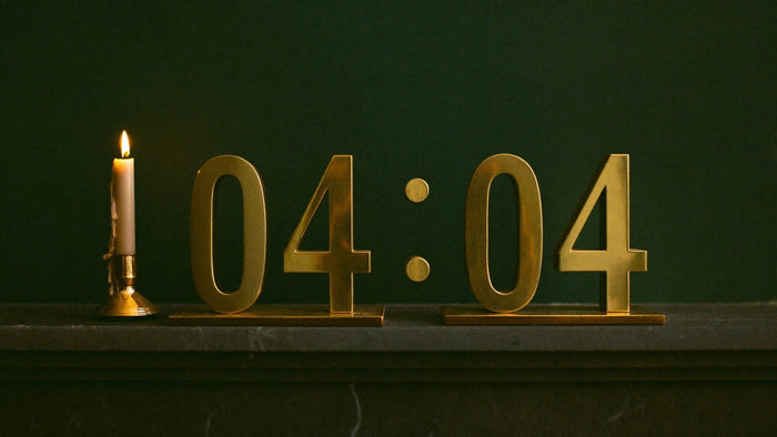 Brass numerals reading 04:04 on a dark stone mantel beside a lit taper candle against a dark green wall