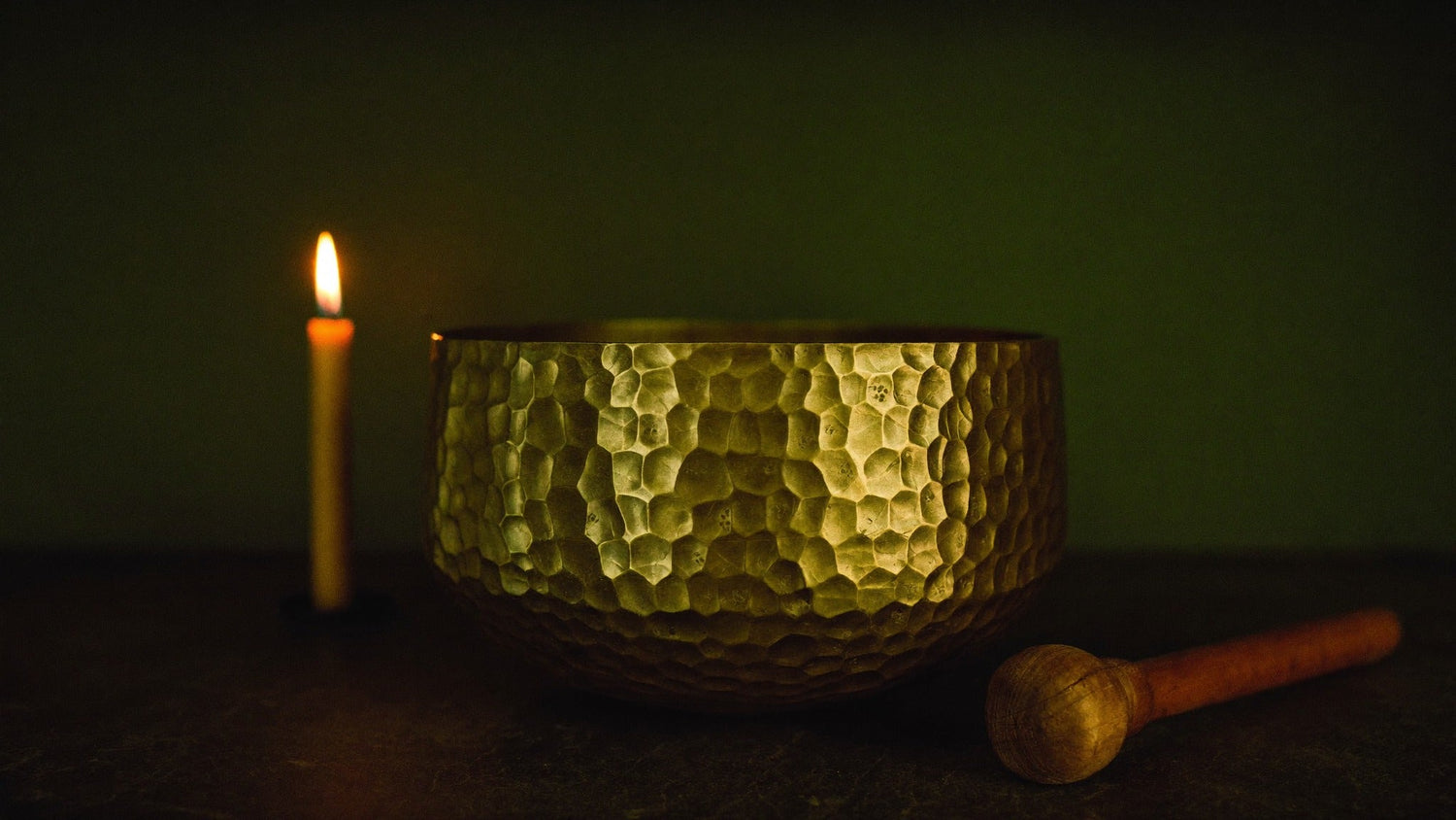 Hammered bronze singing bowl and wooden mallet lit by a single candle against a dark green wall