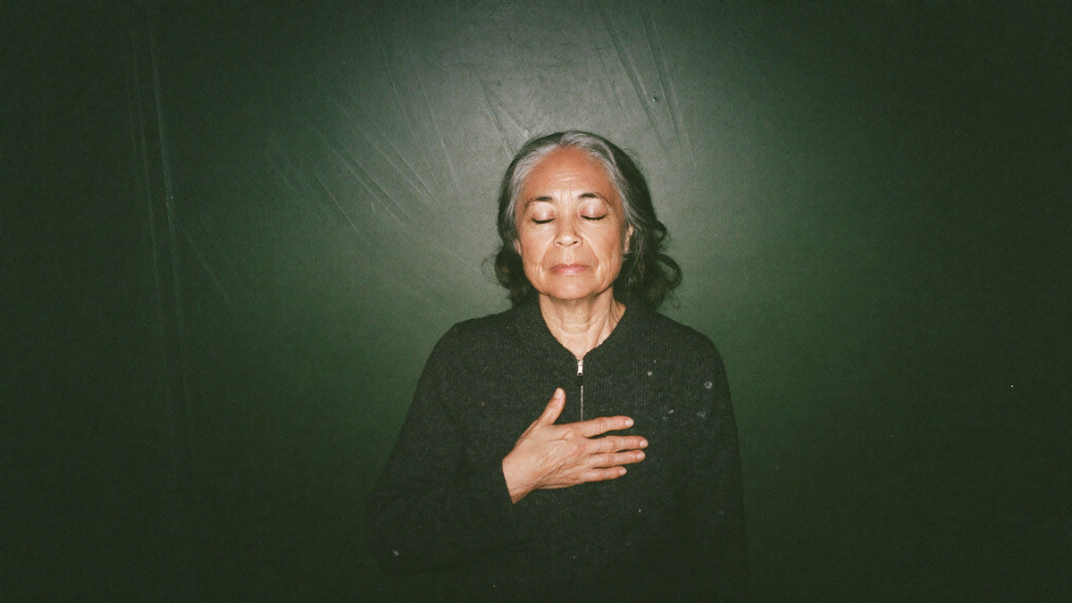 A woman with her eyes closed and one hand resting flat over her heart against a dark green wall