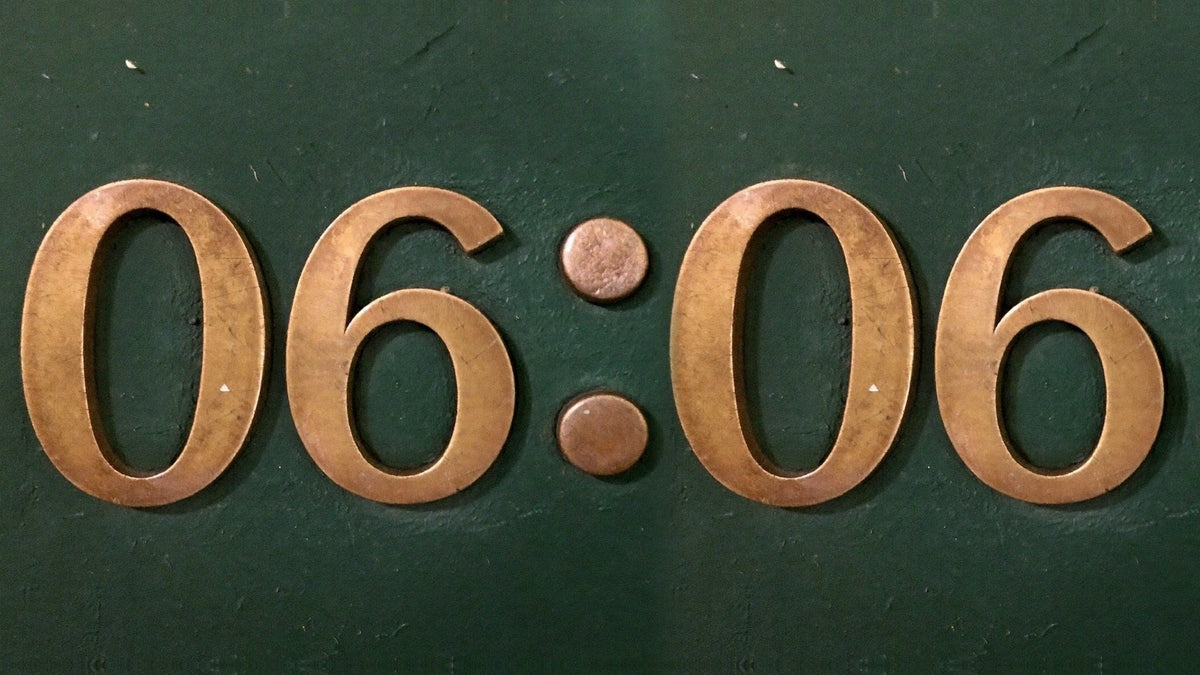 Brass numerals reading 06:06 mounted on a dark green painted wall, warm light on the metal