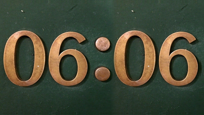 Brass numerals reading 06:06 mounted on a dark green painted wall, warm light on the metal
