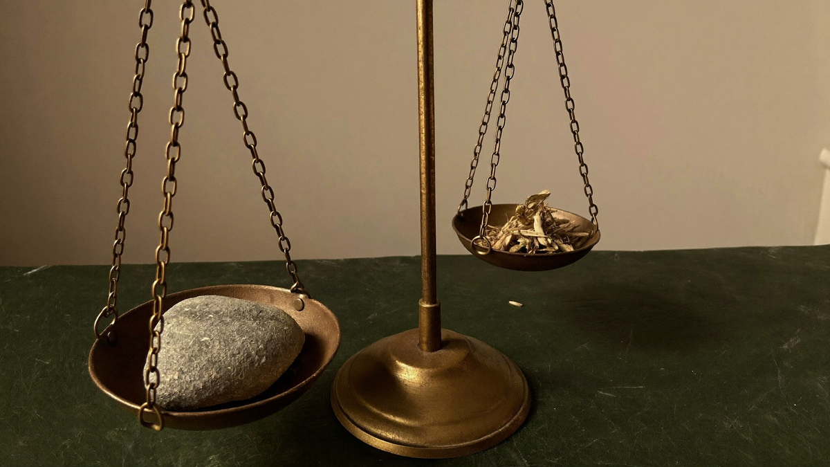 An antique brass balance scale on dark slate, one pan holding a stone and tipping below the other
