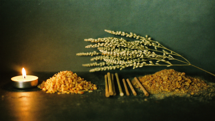 Golden myrrh resin, dried cypress sprigs and incense sticks lit by a single candle on dark stone