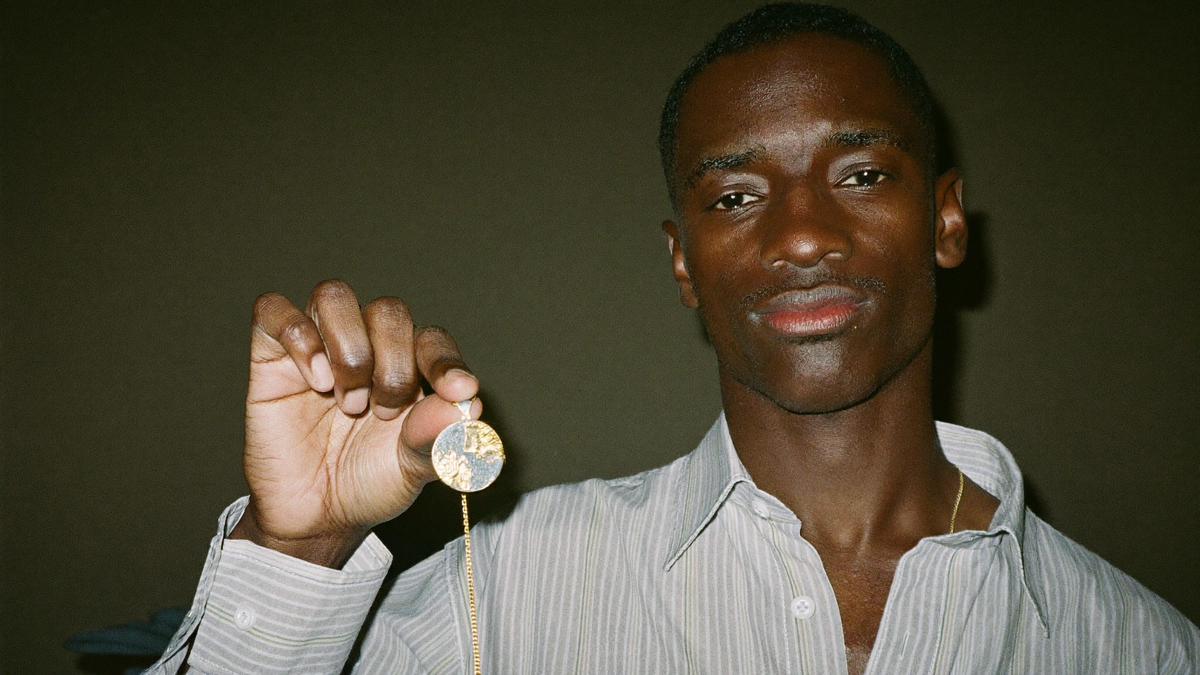 Young man holding up a small round gold pendant on a fine chain against a dark wall