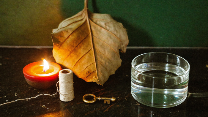 Lit candle, dried leaf, spool of thread, brass key and a bowl of water set out on a dark stone surface