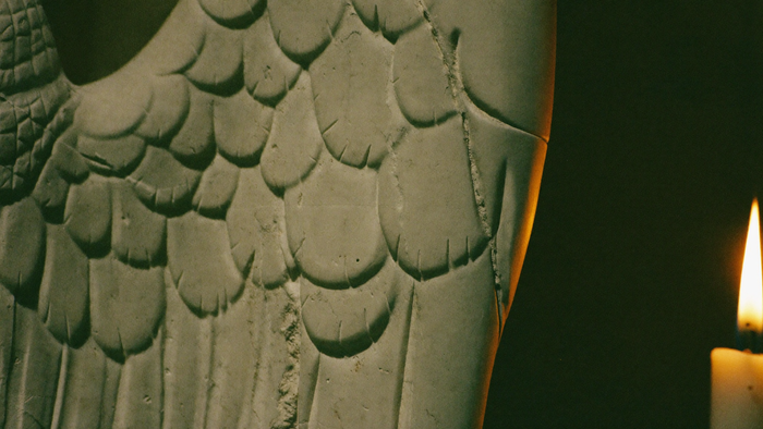 Close view of a carved stone angel wing, feathers cut into weathered pale stone, lit by candlelight