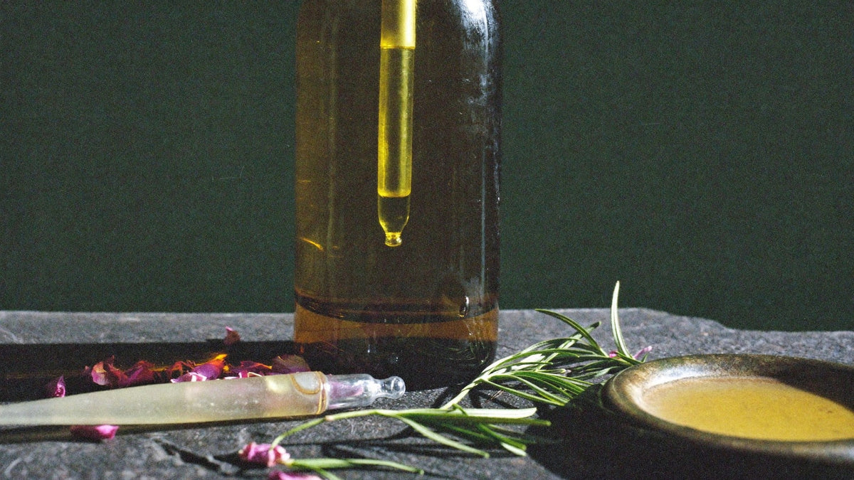 Amber glass oil bottle with a dropper, dried rose petals and rosemary on dark stone