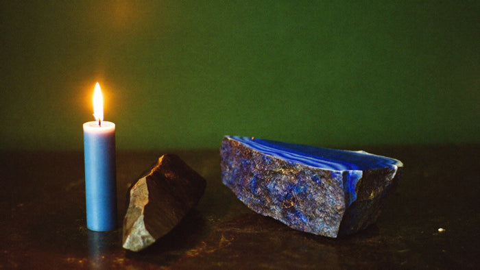 A lit blue candle beside a raw stone and a deep blue lapis slab on a dark surface