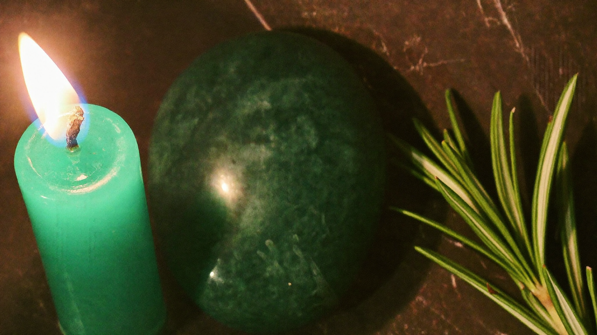 Green candle burning beside a polished green stone and a rosemary sprig on dark stone