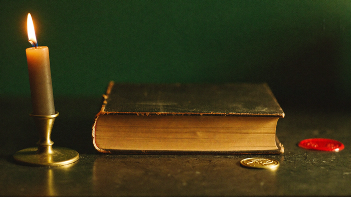 An old leather-bound book with gilded edges beside a brass candlestick, a red wax seal and a gold seal stamp on dark stone
