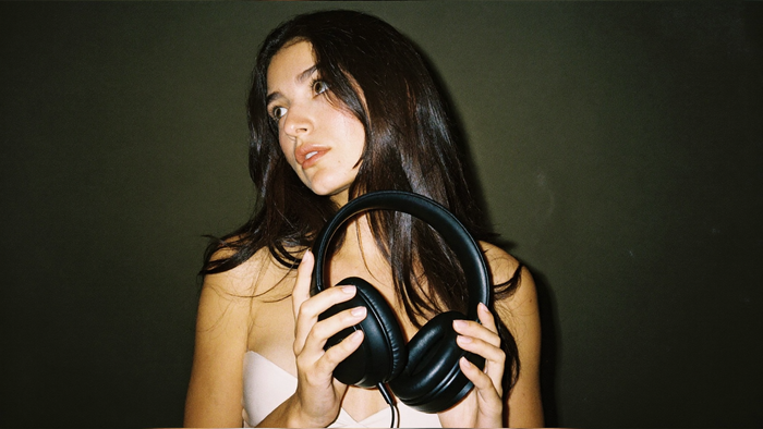 A young woman holding a pair of black over-ear headphones against a dark green wall, lit by direct flash