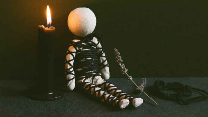 Linen poppet doll bound in black cord beside a burning black candle and a dried lavender sprig on dark slate