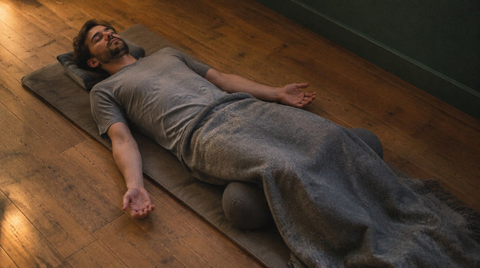Man lying on his back on a floor mat with eyes closed, palms up and a wool blanket over his legs, doing a body scan
