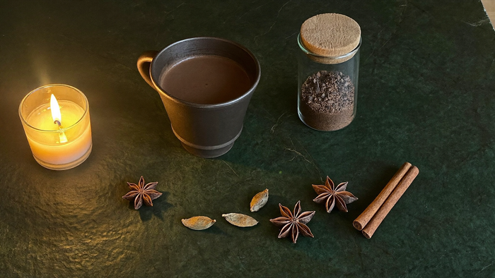 Dark cup of ceremonial cacao beside a lit candle, cacao nibs, star anise and cinnamon on green stone