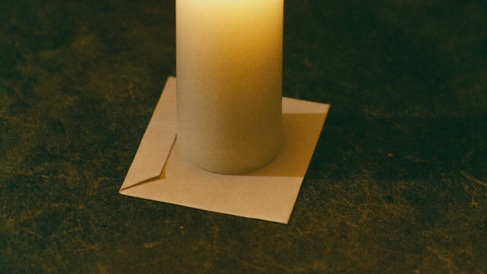 The base of a single white candle standing on a folded slip of paper on dark stone, lit warmly from the left