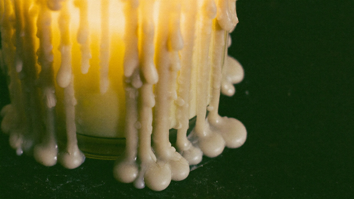 Pale candle wax hardened into thick drips down the side of a glowing glass candle on dark stone