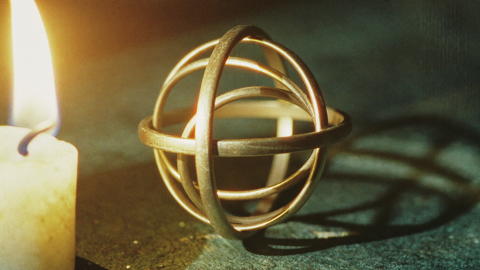 Polished brass armillary sphere lit by a single candle flame on dark stone with a soft shadow